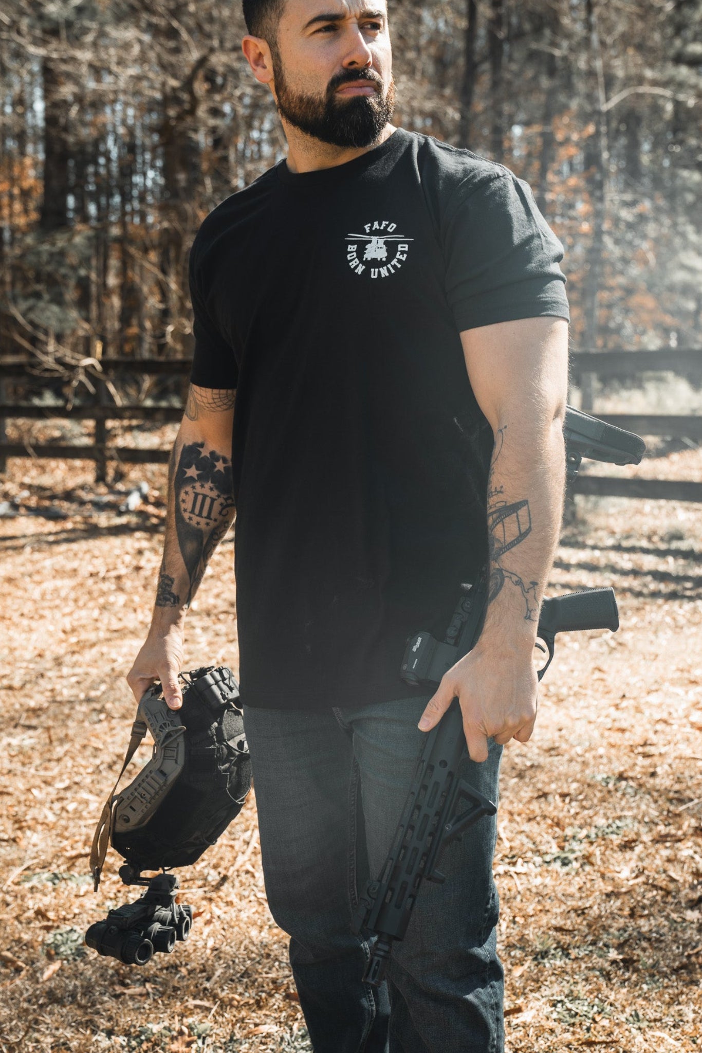 A bearded man stands outdoors on dry grass, holding a helmet and the FAFO - Maduro firearm by Born United Private Label. He wears a black Limited Design shirt, jeans, and displays tattoos on both arms. Trees and a wooden fence are behind him.