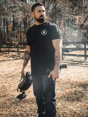 A bearded man stands outdoors on dry grass, holding a helmet and the FAFO - Maduro firearm by Born United Private Label. He wears a black Limited Design shirt, jeans, and displays tattoos on both arms. Trees and a wooden fence are behind him.
