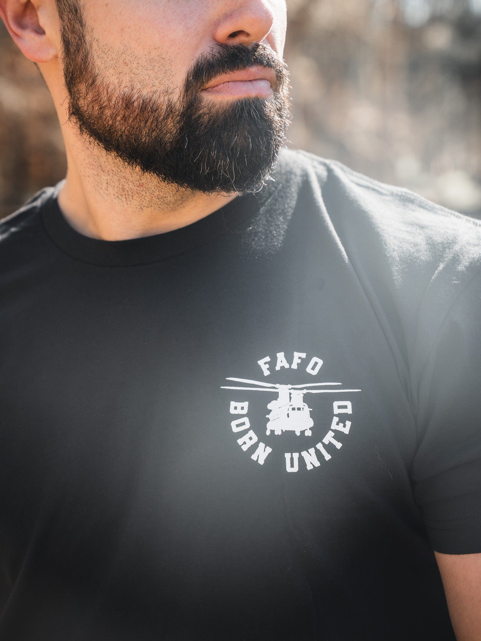 A bearded man wears the FAFO – Maduro black tee by Born United Private Label, featuring a white logo and small vehicle graphic. This limited design is seen outdoors with sunlight highlighting its bold style.