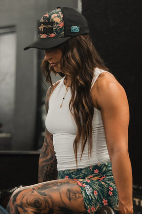 A woman with long hair sits indoors, possibly at a gym, showing off her tattooed arm. She wears a breathable Aloha AR Hydro - Snapback Hat by Tango Charlie Apparel, a white sleeveless top, and floral-patterned shorts.
