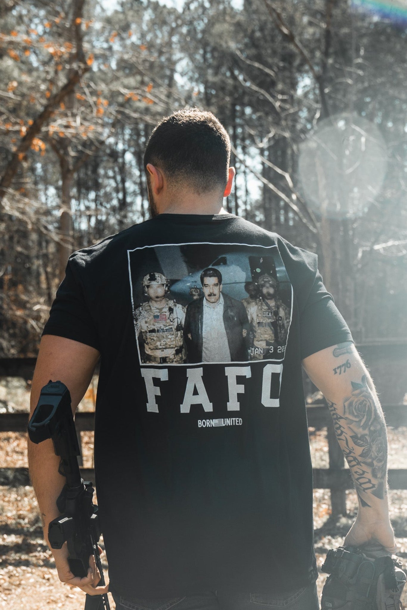 A man stands outdoors with his back to the camera, holding a firearm and helmet. He wears the limited edition Born United Private Label "FAFO - Maduro" black shirt as sunlight filters through the trees behind him.