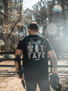 A man stands outdoors with his back to the camera, holding a firearm and helmet. He wears the limited edition Born United Private Label "FAFO - Maduro" black shirt as sunlight filters through the trees behind him.