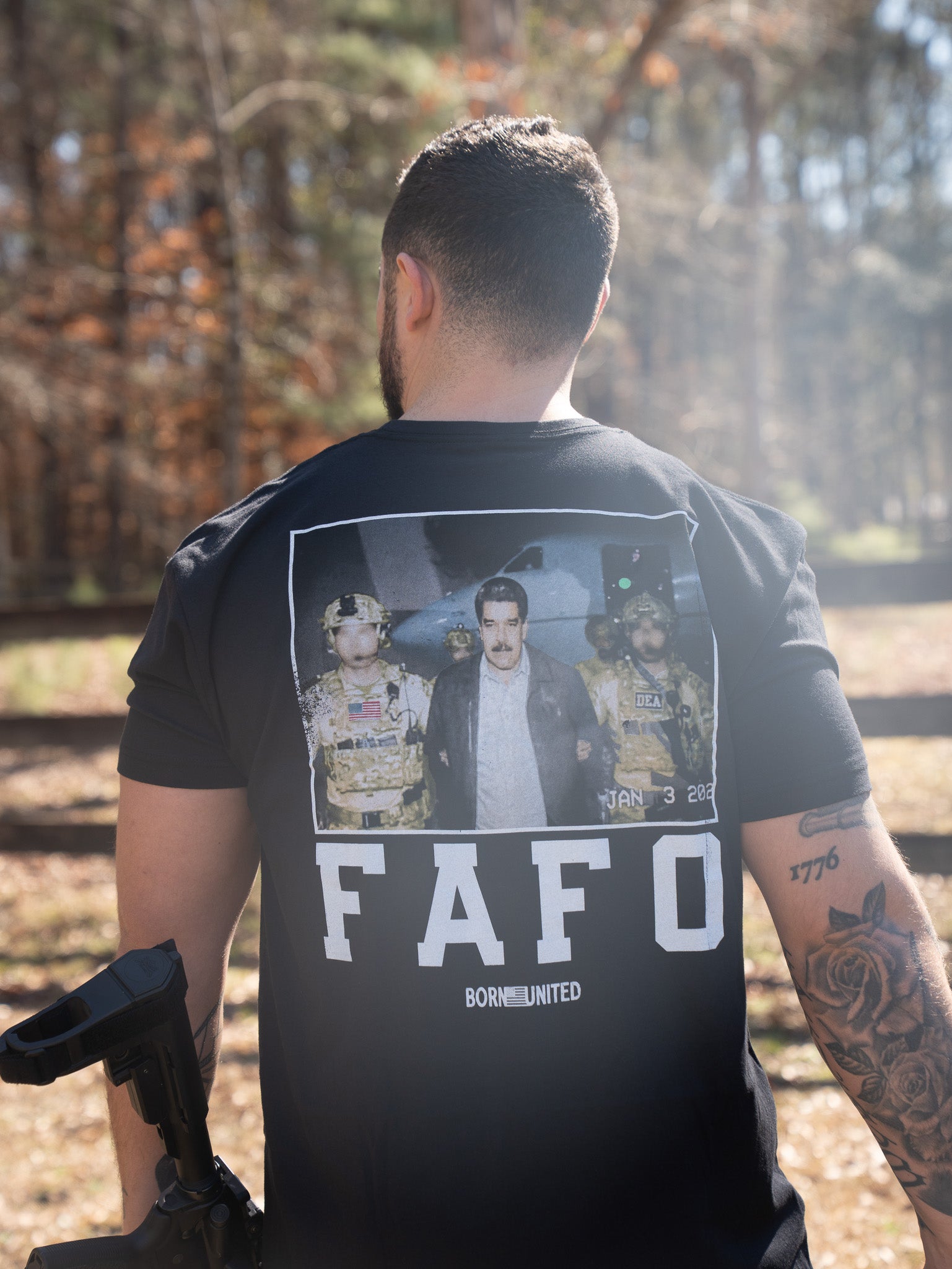 A man stands outdoors with his back to the camera, wearing the Born United Private Label FAFO - Maduro T-shirt, which features a photo of an arrest scene and bold FAFO and BORN UNITED text. Sunlight filters through the trees behind him.