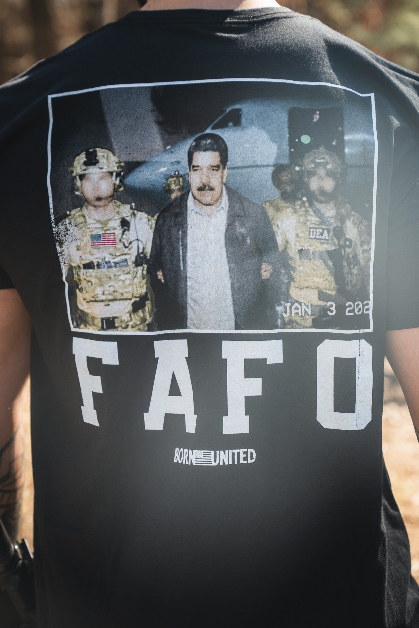 A person wears the FAFO - Maduro black T-shirt by Born United Private Label, featuring a graphic of a man escorted by armed officers and "FAFO" and "BORN UNITED" printed below, mimicking a dramatic news photo or arrest scene.
