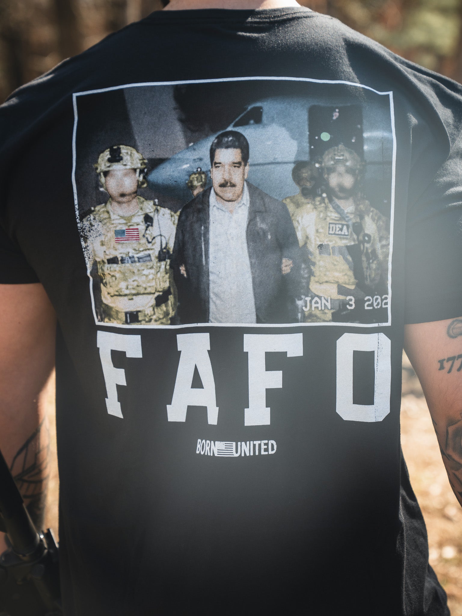 A person wears the FAFO - Maduro black T-shirt by Born United Private Label, featuring a graphic of a man escorted by armed officers and "FAFO" and "BORN UNITED" printed below, mimicking a dramatic news photo or arrest scene.