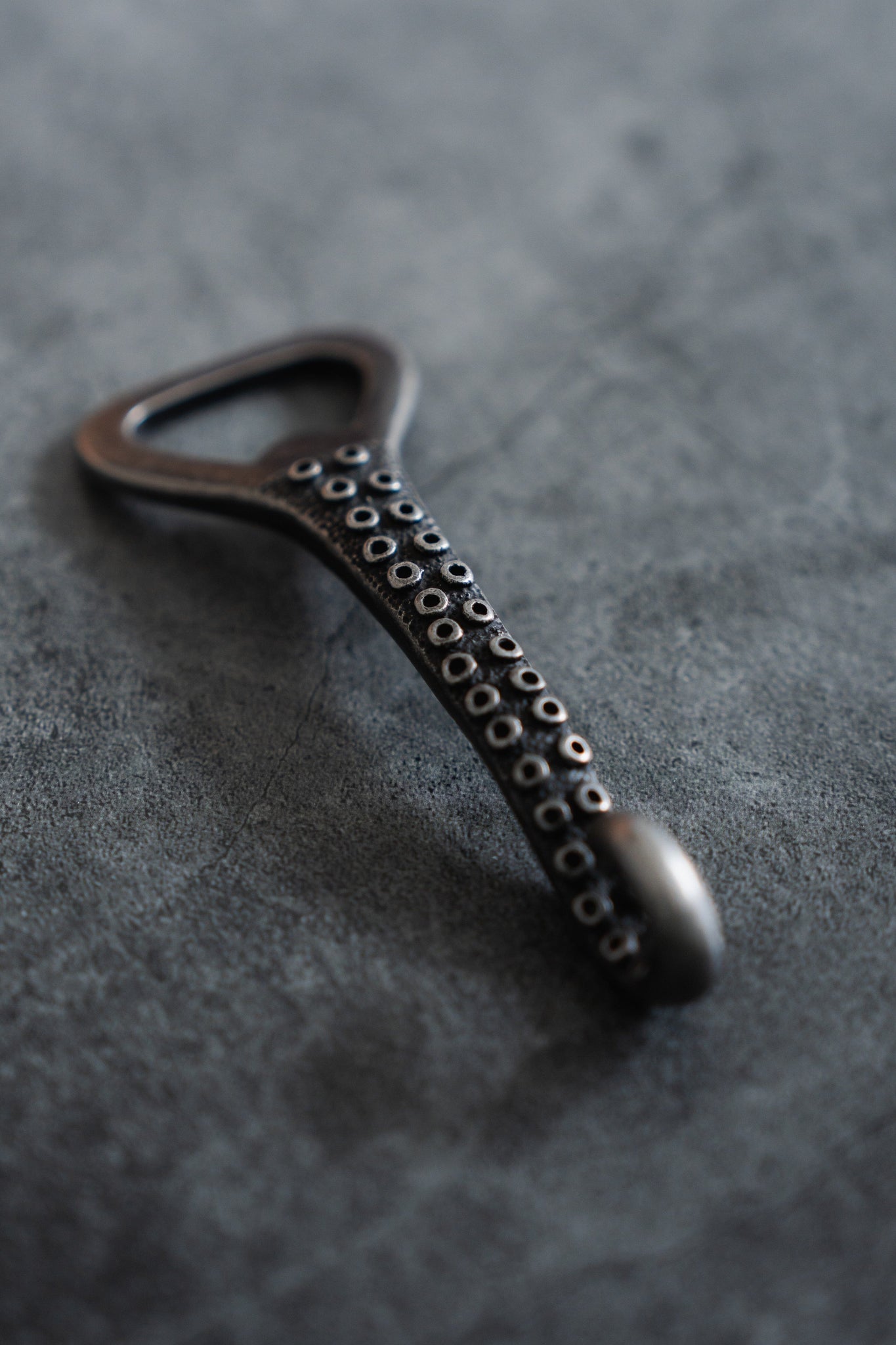 The Kraken Bottle Opener by Relentless Betrayal is a USA-designed stainless steel bottle opener shaped like an octopus tentacle with suction cup details, set against a textured gray background.
