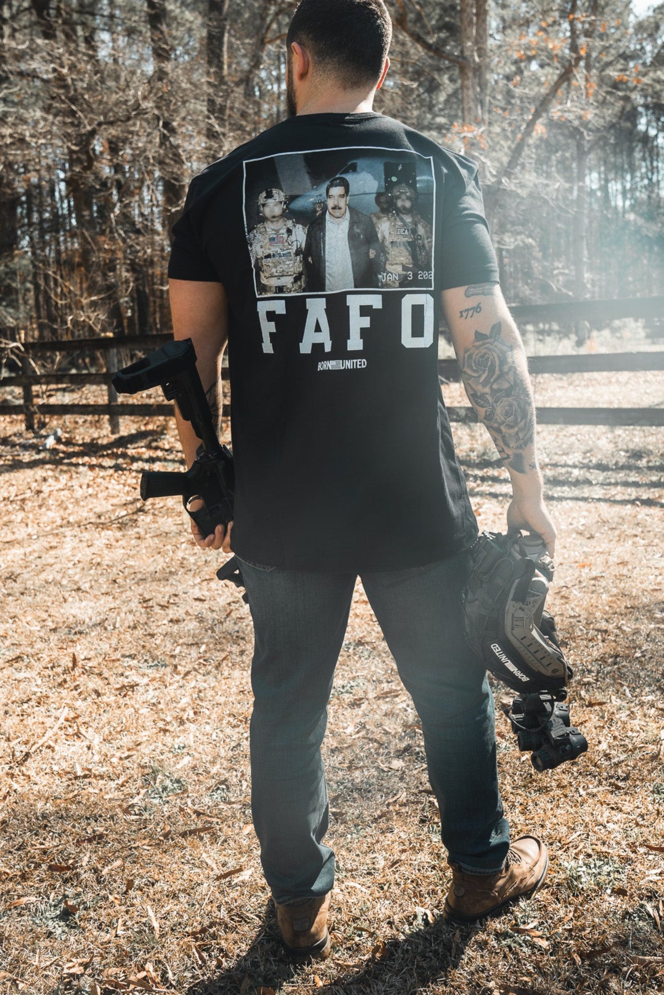 A man stands outdoors in a grassy area, holding a helmet and rifle. He wears the Born United Private Label "FAFO - Maduro" shirt with an image on the back. Tall trees and a wooden fence are visible in the background.