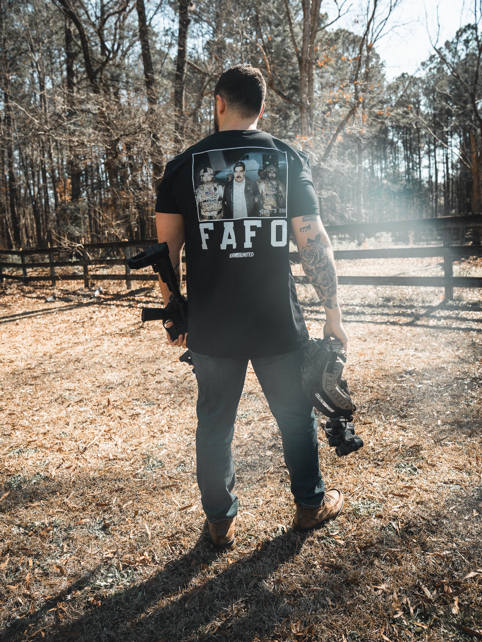 A man stands outdoors in a grassy area, holding a helmet and rifle. He wears the Born United Private Label "FAFO - Maduro" shirt with an image on the back. Tall trees and a wooden fence are visible in the background.