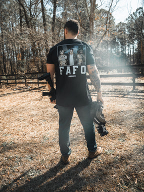 A man stands outdoors in a grassy area, holding a helmet and rifle. He wears the Born United Private Label "FAFO - Maduro" shirt with an image on the back. Tall trees and a wooden fence are visible in the background.