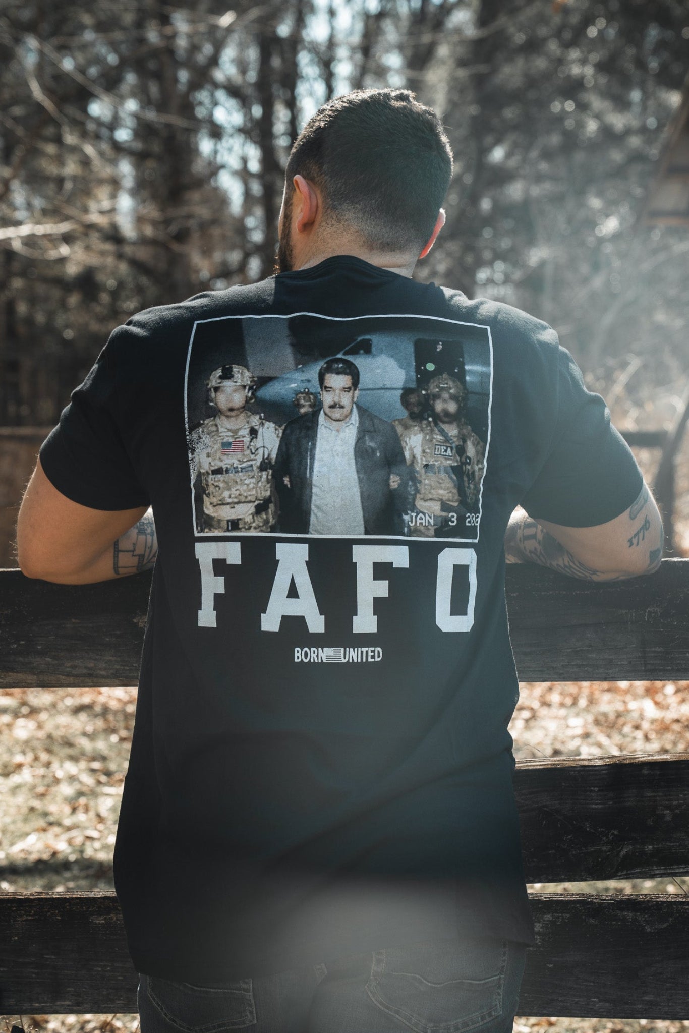 A tattooed man stands outdoors by a fence, wearing a Born United Private Label FAFO - Maduro black t-shirt with three people and a “FAFO BORN UNITED” graphic on the back. Trees and a wooden building are visible behind him.