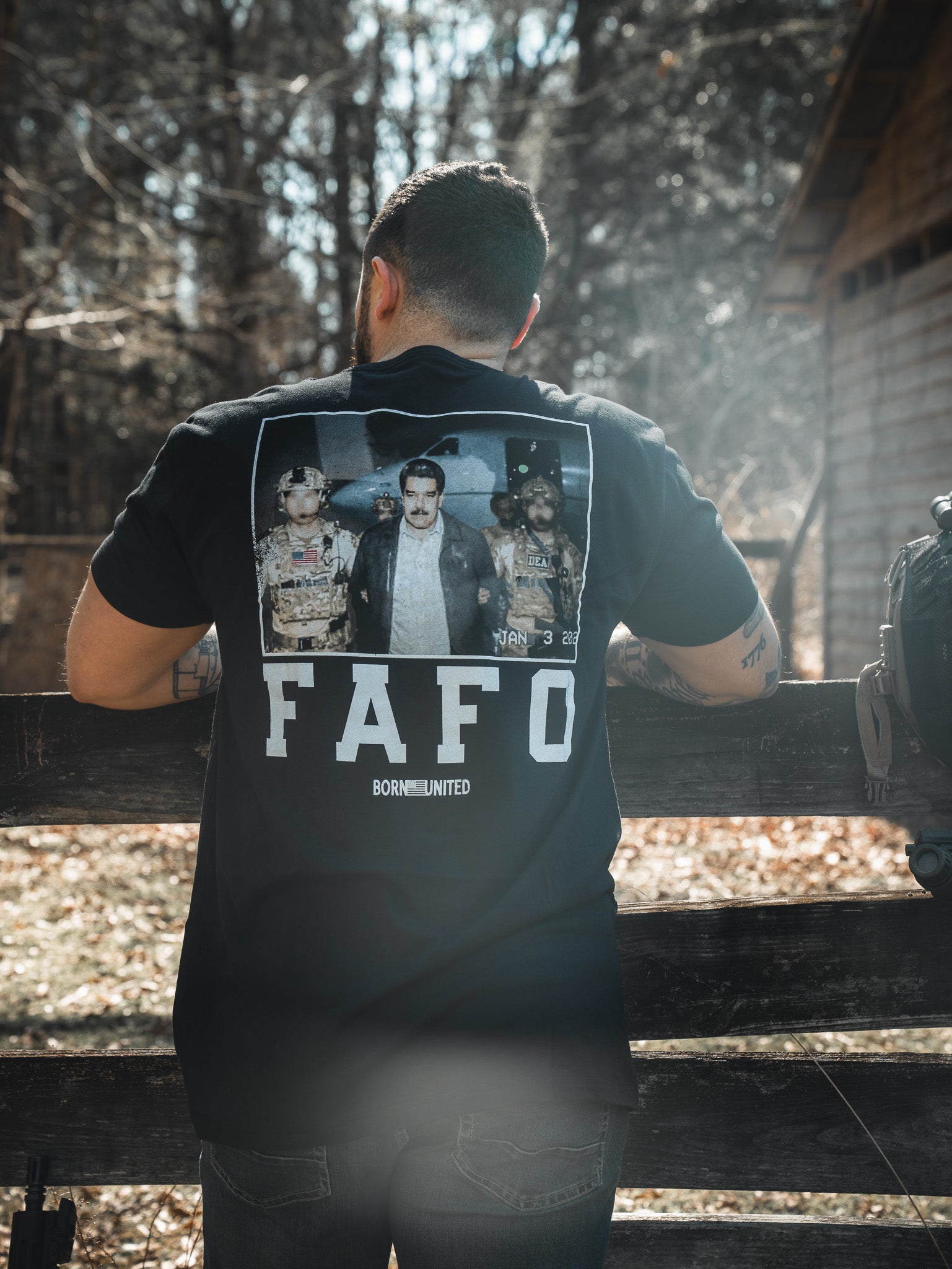 A tattooed man stands outdoors by a fence, wearing a Born United Private Label FAFO - Maduro black t-shirt with three people and a “FAFO BORN UNITED” graphic on the back. Trees and a wooden building are visible behind him.