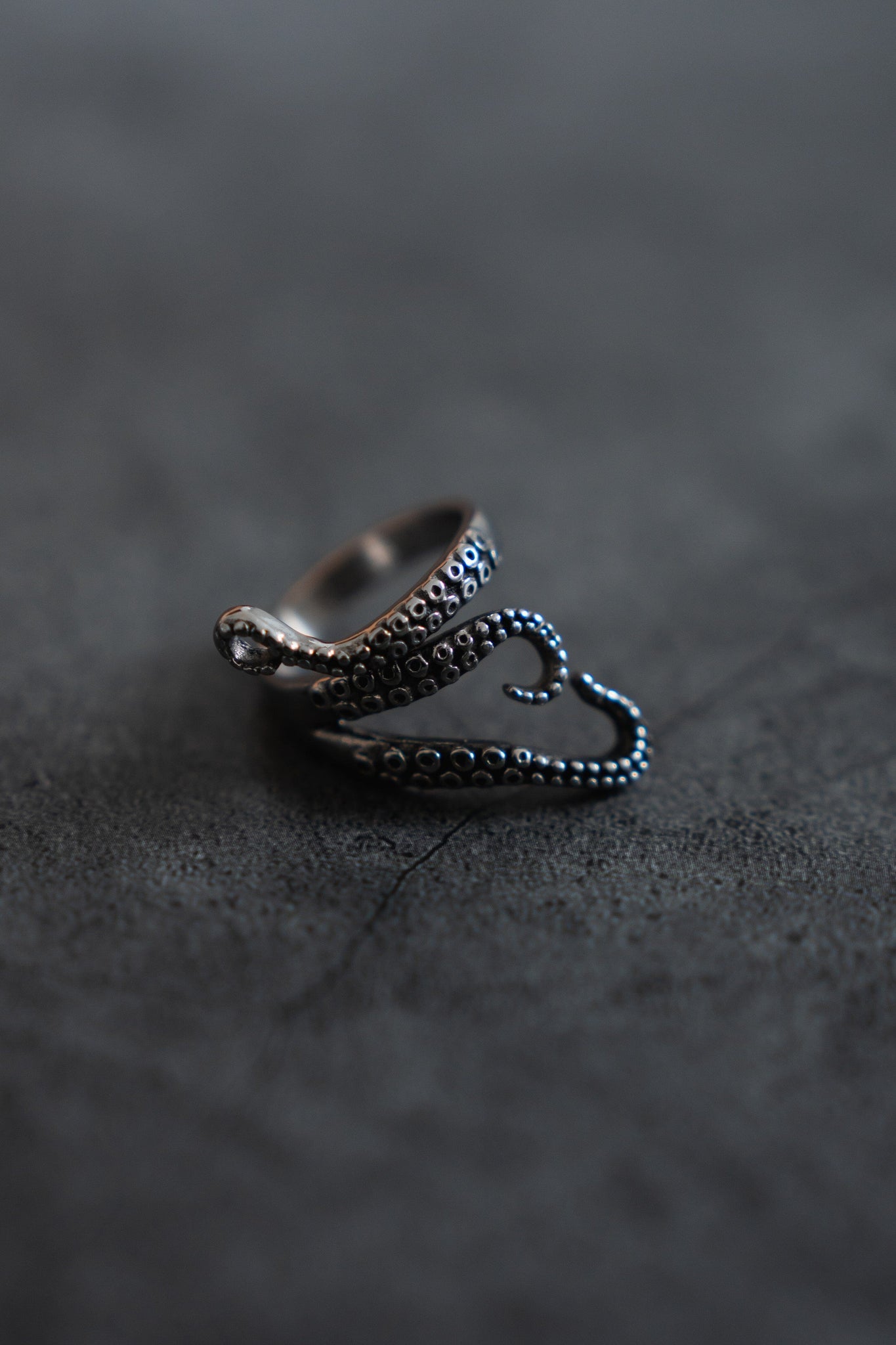The Kraken Stainless Steel ring by Relentless Betrayal features a meticulously designed, silver-like finish that resembles a coiled octopus tentacle with detailed texture, resting on a dark surface. Its intricate design echoes the mythical krakens allure.
