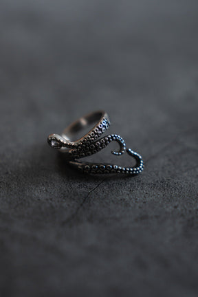 The Kraken Stainless Steel ring by Relentless Betrayal features a meticulously designed, silver-like finish that resembles a coiled octopus tentacle with detailed texture, resting on a dark surface. Its intricate design echoes the mythical krakens allure.
