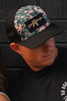 A man in a Tango Charlie Apparel Aloha AR Hydro Snapback hat with a floral and embroidered gun motif looks down. Against a dark brick wall, his black t-shirt complements this water-resistant hat, merging function with flair.