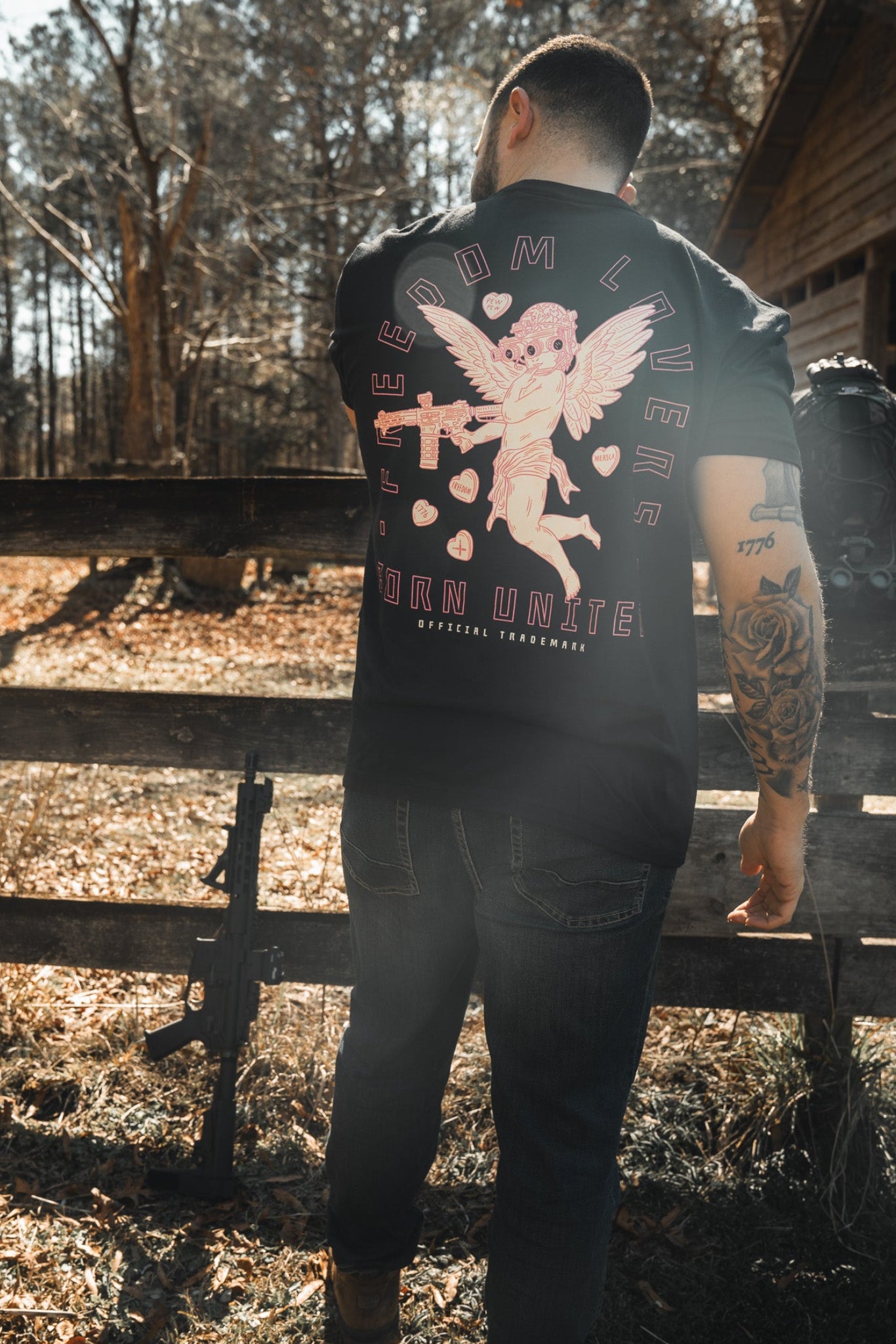 A tattooed man stands outdoors by a wooden fence, sporting Born United Private Label’s edgy "Cupid’s Shot" Valentine’s tee with a winged skeleton, holding a camera and posing with a nearby rifle—the perfect tactical cupid vibe.