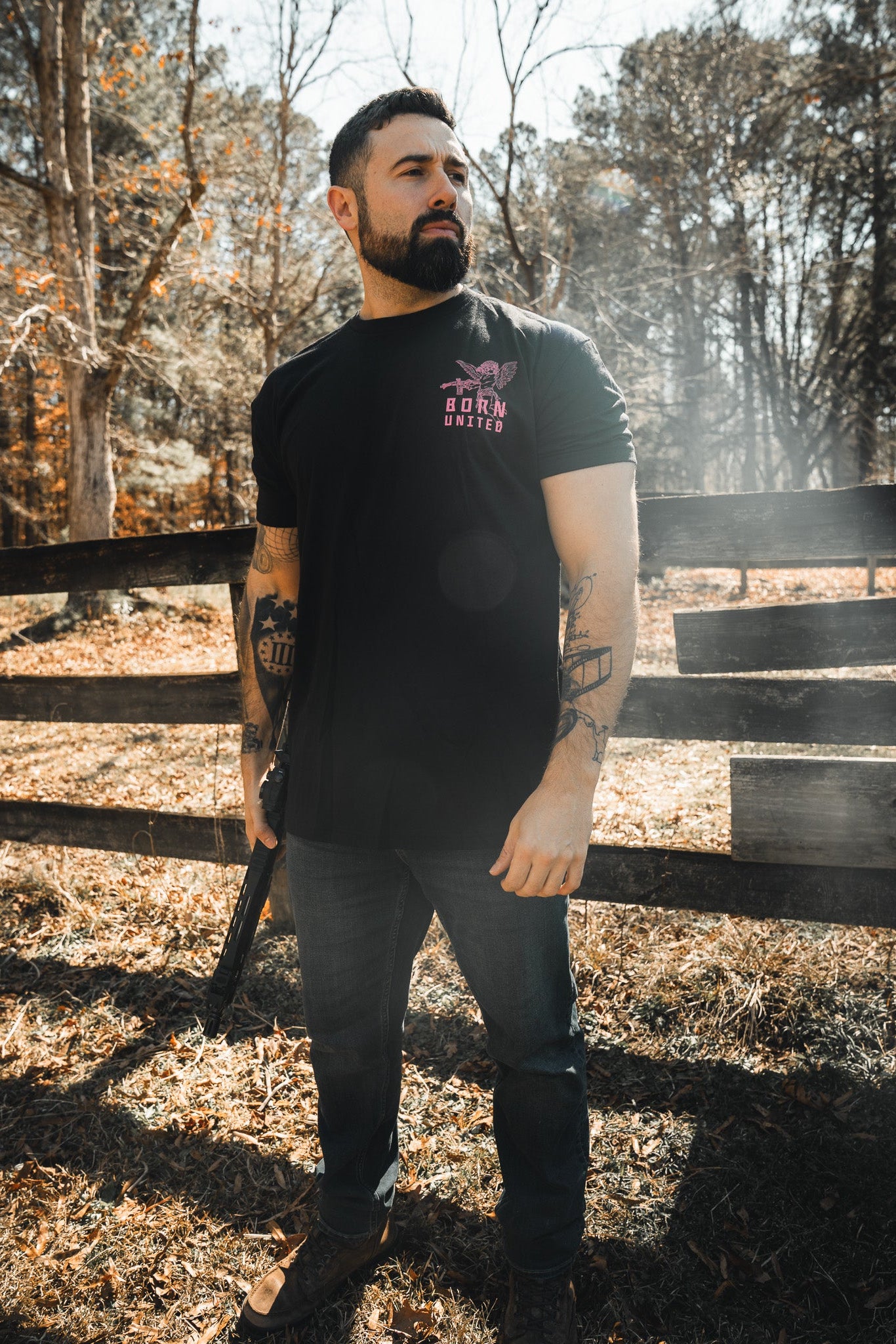 A tattooed bearded man stands outdoors by a wooden fence, wearing the “Cupid’s Shot” Valentine's Day shirt from Born United Private Label. Dressed in jeans and boots, he holds an object at his side as sunlight filters through the trees.