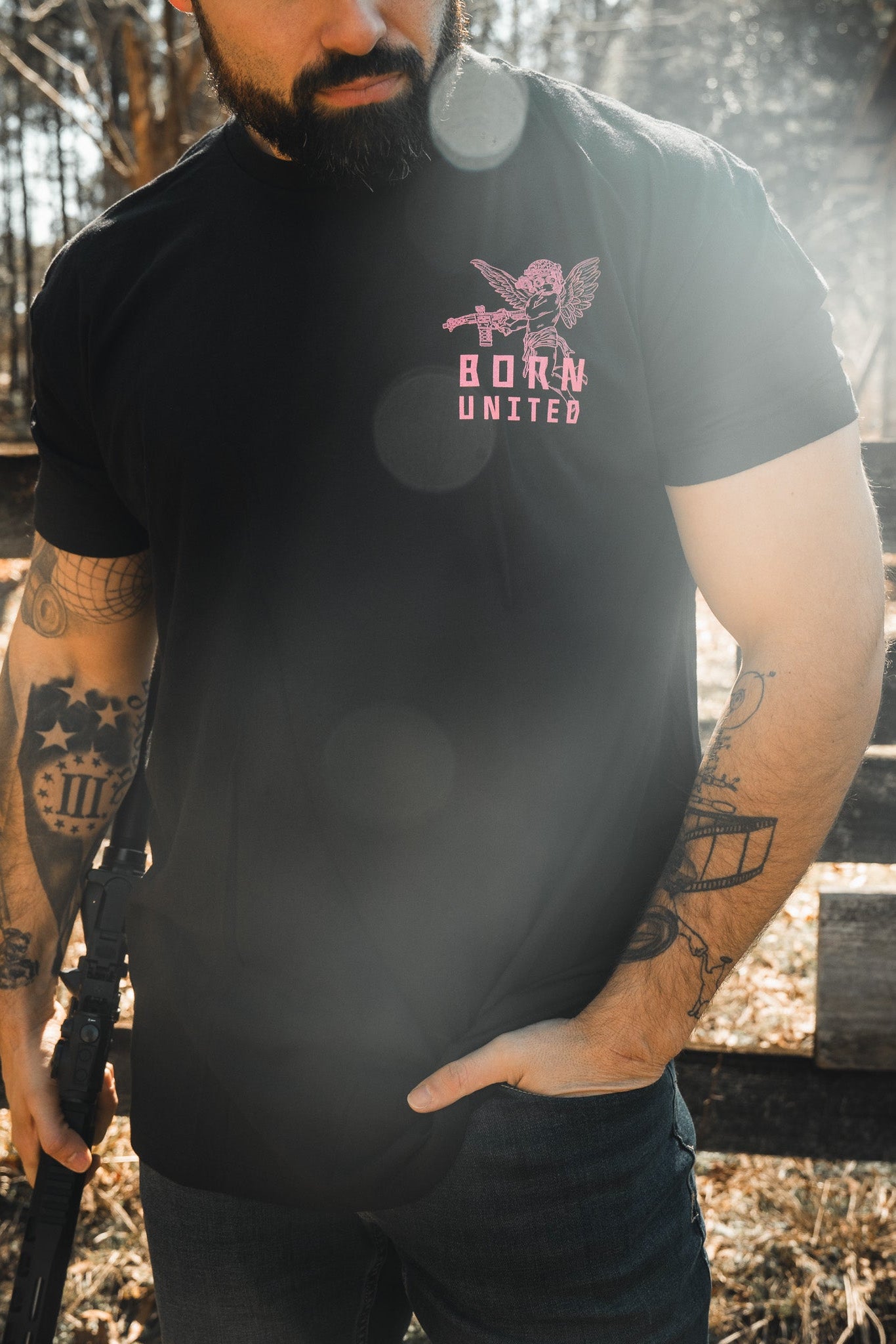 A tattooed bearded man stands outdoors holding a rifle, wearing the "Cupid’s Shot" tee by Born United Private Label—a bold Valentine’s Day shirt with a pink tactical cupid and “BORN UNITED” text. Lens flares catch in the sunlight.