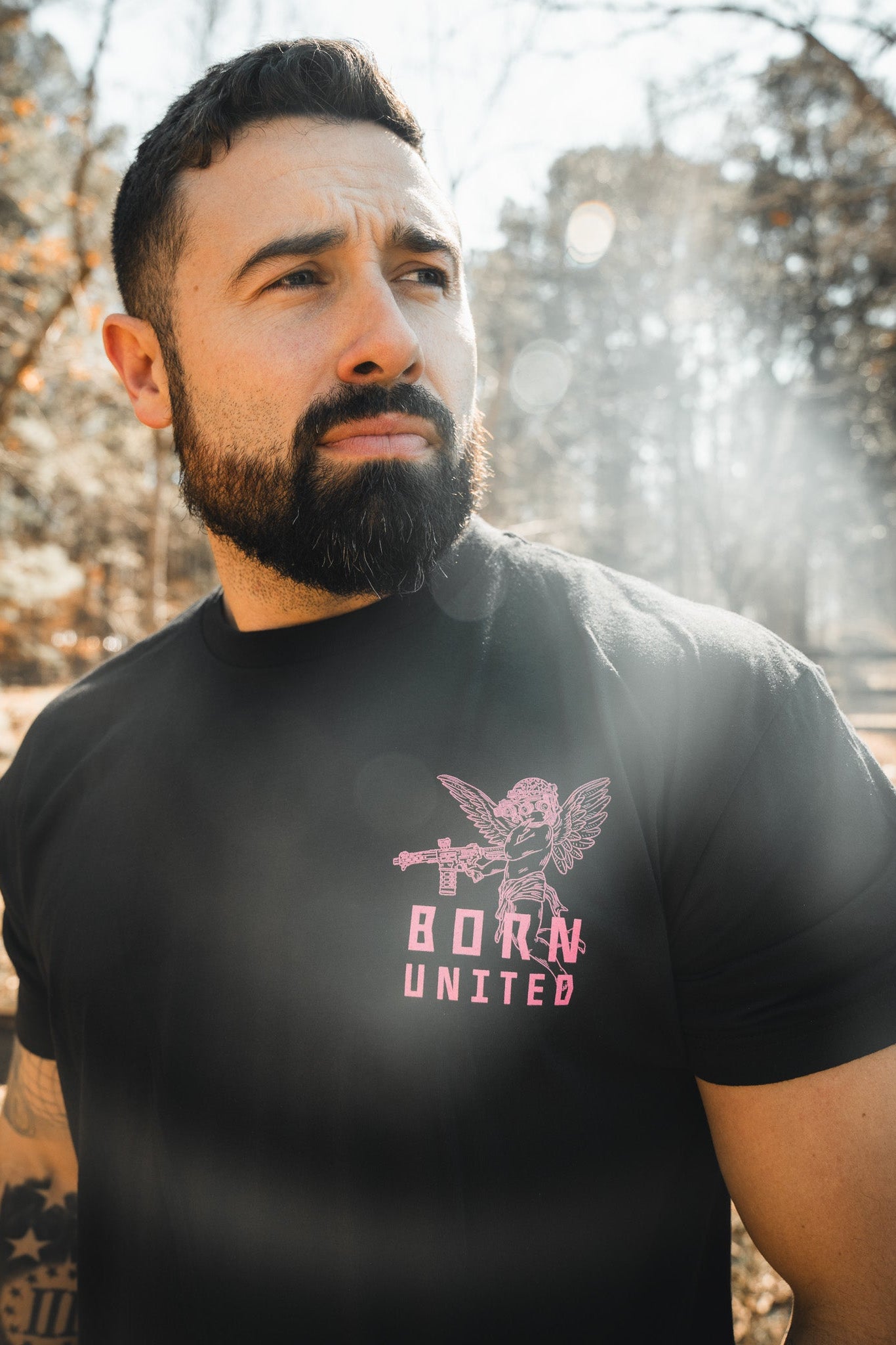 A bearded man stands outdoors in sunlight, wearing the Born United Private Label "Cupid’s Shot" shirt featuring a pink tactical cupid graphic. Trees and a wooden fence appear in the background.