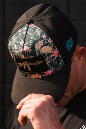 A man wears the Tango Charlie Apparel Aloha AR Hydro snapback hat, featuring a black cap with a floral and skull design, a gold gun graphic, and a blue logo on the side. The water-resistant hat offers breathability for comfort as he tilts his head forward, touching its brim with his right hand.