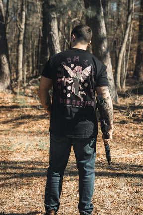 A man stands in a sunlit forest, facing away as he holds a rifle. He wears jeans and a Born United Private Label "Cupid’s Shot" Valentine’s Day shirt with a bold winged graphic and text on the back.
