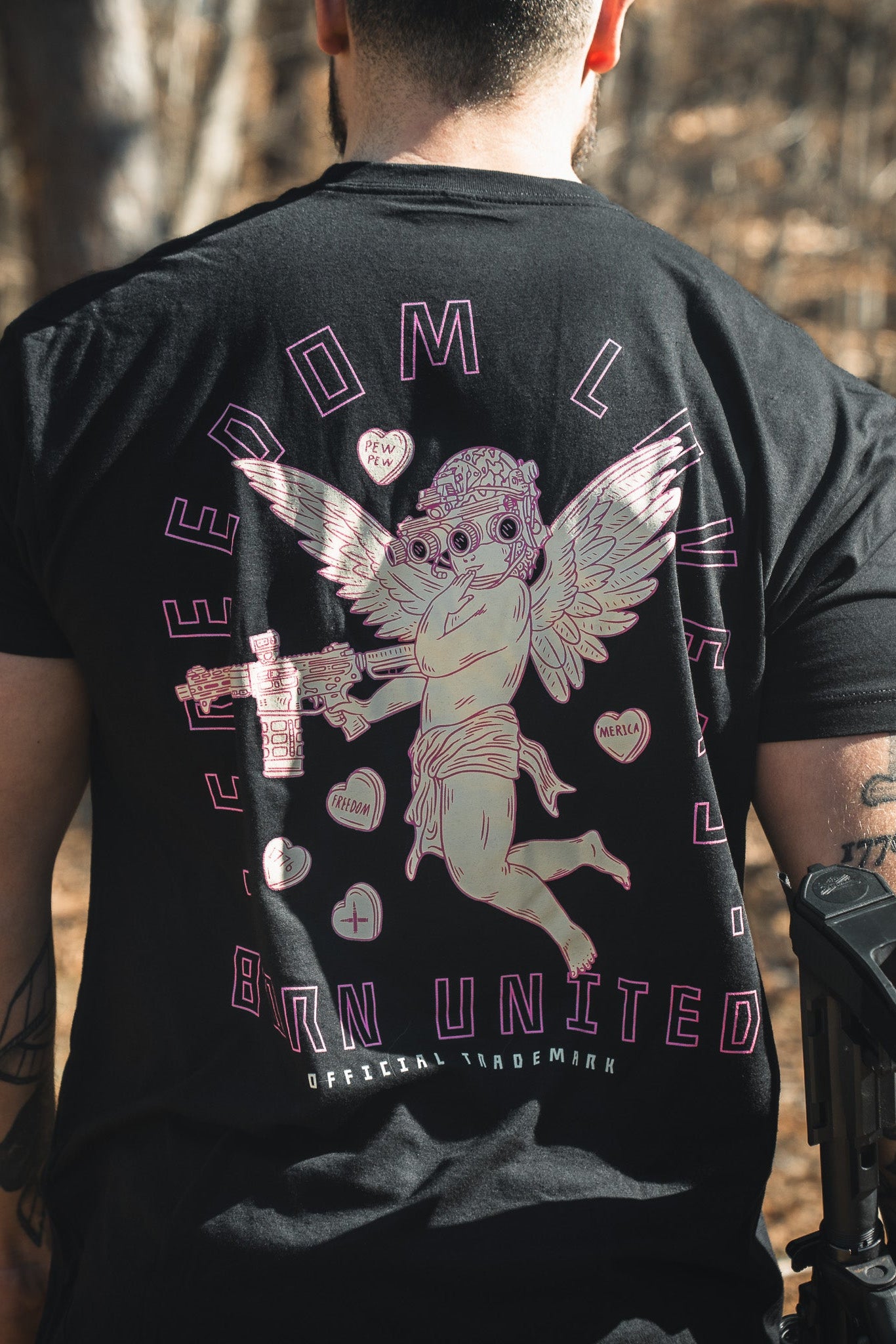 Outdoors, a person (back turned) wears the Born United Private Label “Cupid’s Shot” tee: a black shirt featuring a tactical cherub with gun and drink, surrounded by “FREEDOM LIVES,” “BORN UNITED,” and bold symbols.