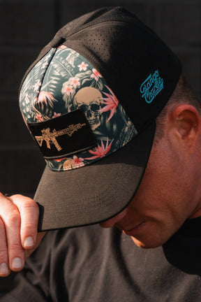 A person dons a Tango Charlie Apparel Aloha AR Hydro Snapback Hat in black, flaunting floral and skull designs. The water-resistant cap includes a rifle patch and Guaro Mercado text. Theyre looking down with part of their face obscured.