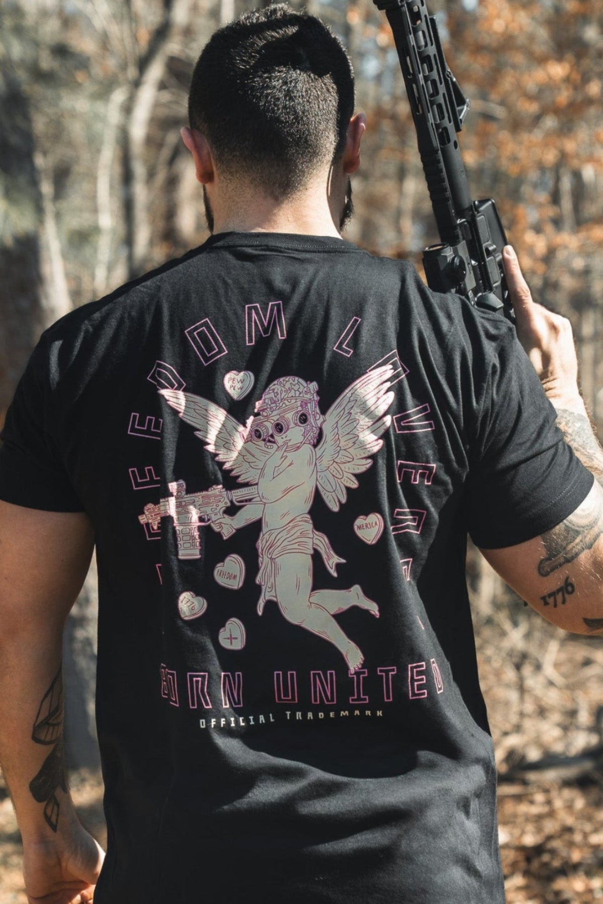 A man with short dark hair stands outdoors holding a rifle over his right shoulder, wearing the Born United Private Label "Cupid’s Shot" t-shirt, featuring an angel graphic and the words FREEDOM LIVES and BORN UNITED on the back.