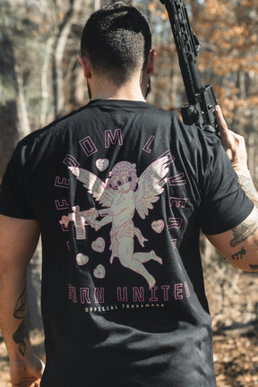 A man with short dark hair stands outdoors holding a rifle over his right shoulder, wearing the Born United Private Label "Cupid’s Shot" t-shirt, featuring an angel graphic and the words FREEDOM LIVES and BORN UNITED on the back.