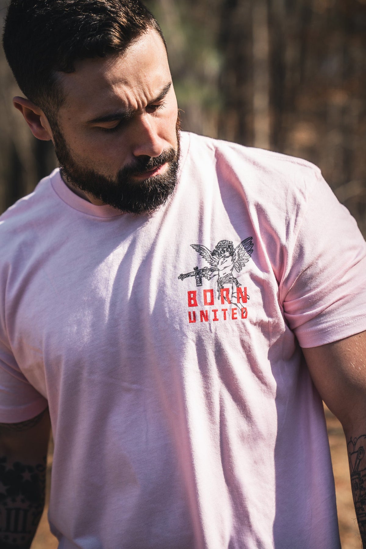 A bearded man stands outdoors among trees, wearing a light pink Cupid’s Shot V2 tee by Born United Private Label, featuring an angel graphic and bold red "BORN UNITED" text, looking down.