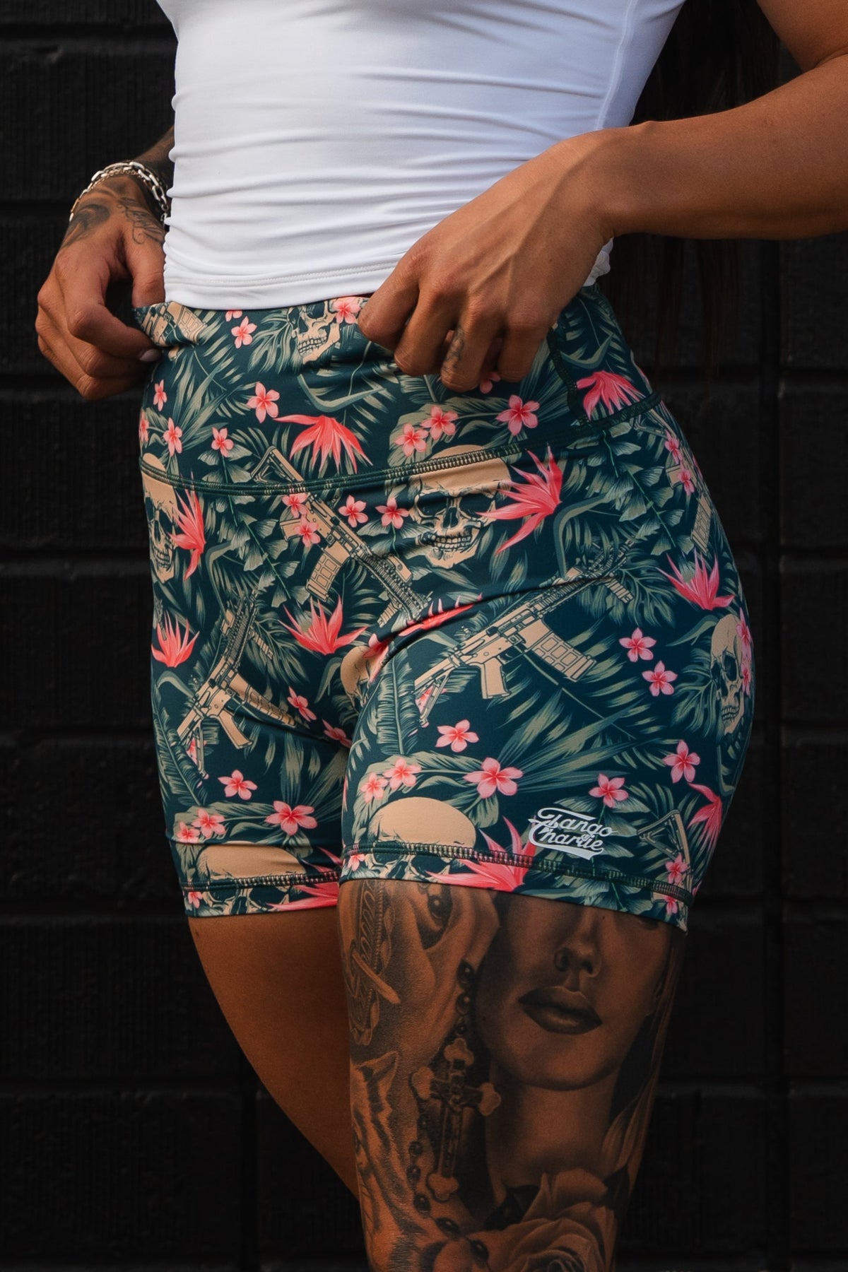 Wearing a white top paired with Tango Charlie Apparels Womens High-Waisted Seamless Front Compression Training Shorts in Aloha AR design, featuring tropical and skull patterns, their thigh tattoos pop against a black textured wall, creating an empowering vibe.