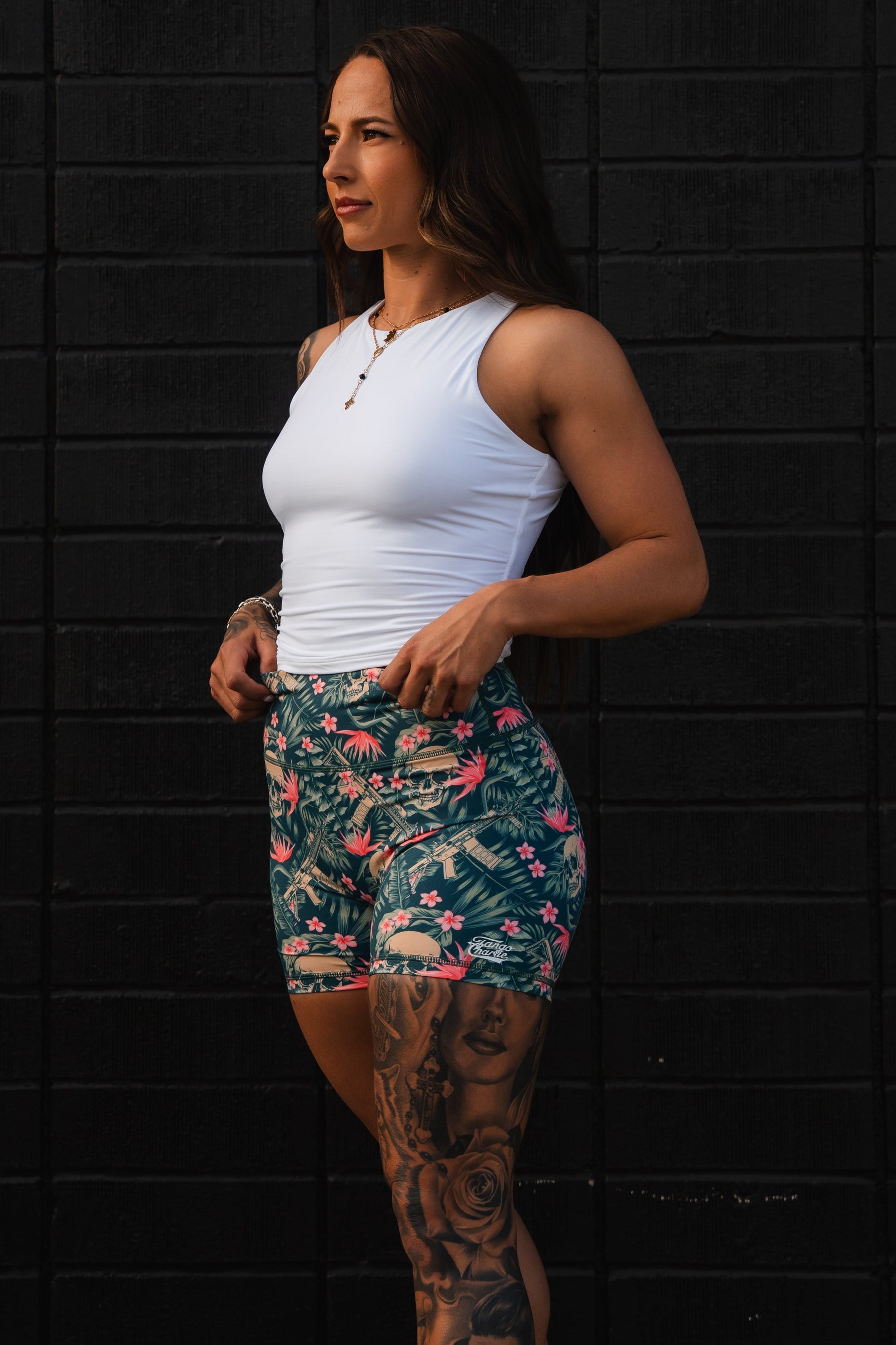 A woman in a white tank top and Tango Charlie Apparel Women's High-Waisted Seamless Front Compression Training Shorts | Aloha AR stands before a black brick wall.
