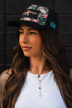 Wearing a black Aloha AR Hydro - Snapback Hat by Tango Charlie Apparel, adorned with tropical designs and colorful patches, the person, with long brown hair in a white top, showcases a necklace featuring cross and crescent moon pendants against a dark, textured wall while looking to the side.