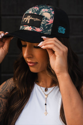 A person with long hair adjusts their Tango Charlie Apparels Aloha AR Hydro Snapback Hat, featuring a floral pattern and front graphic. Theyre in a white top with layered necklaces and cross pendants, standing confidently against a dark backdrop.