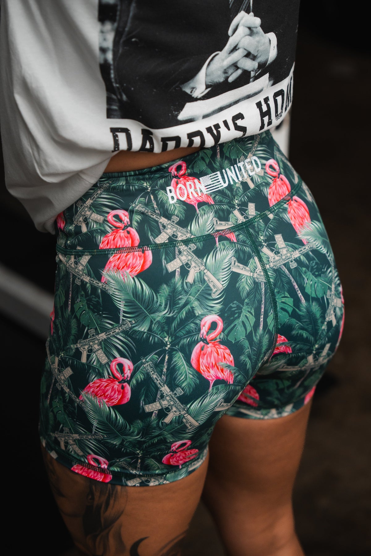 The person wears Womens High-Waisted Seamless Front Compression Training Shorts in a Loaded Flamingo print, made from a breathable fabric by Born United Private Label. A partially visible white t-shirt with BORN UNITED adds to the look against the dark background.