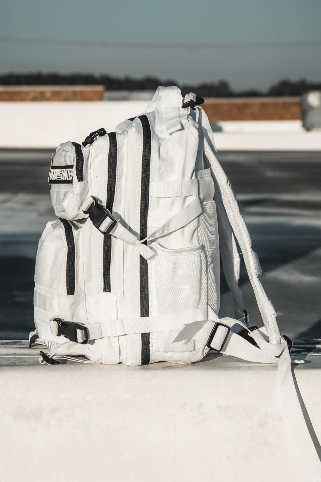 The Born United Private Label Storm Trooper Tac-Pac - 25L, featuring black zippers and straps, sits on a concrete ledge in an urban outdoor setting under clear skies, giving off subtle Star Wars vibes.