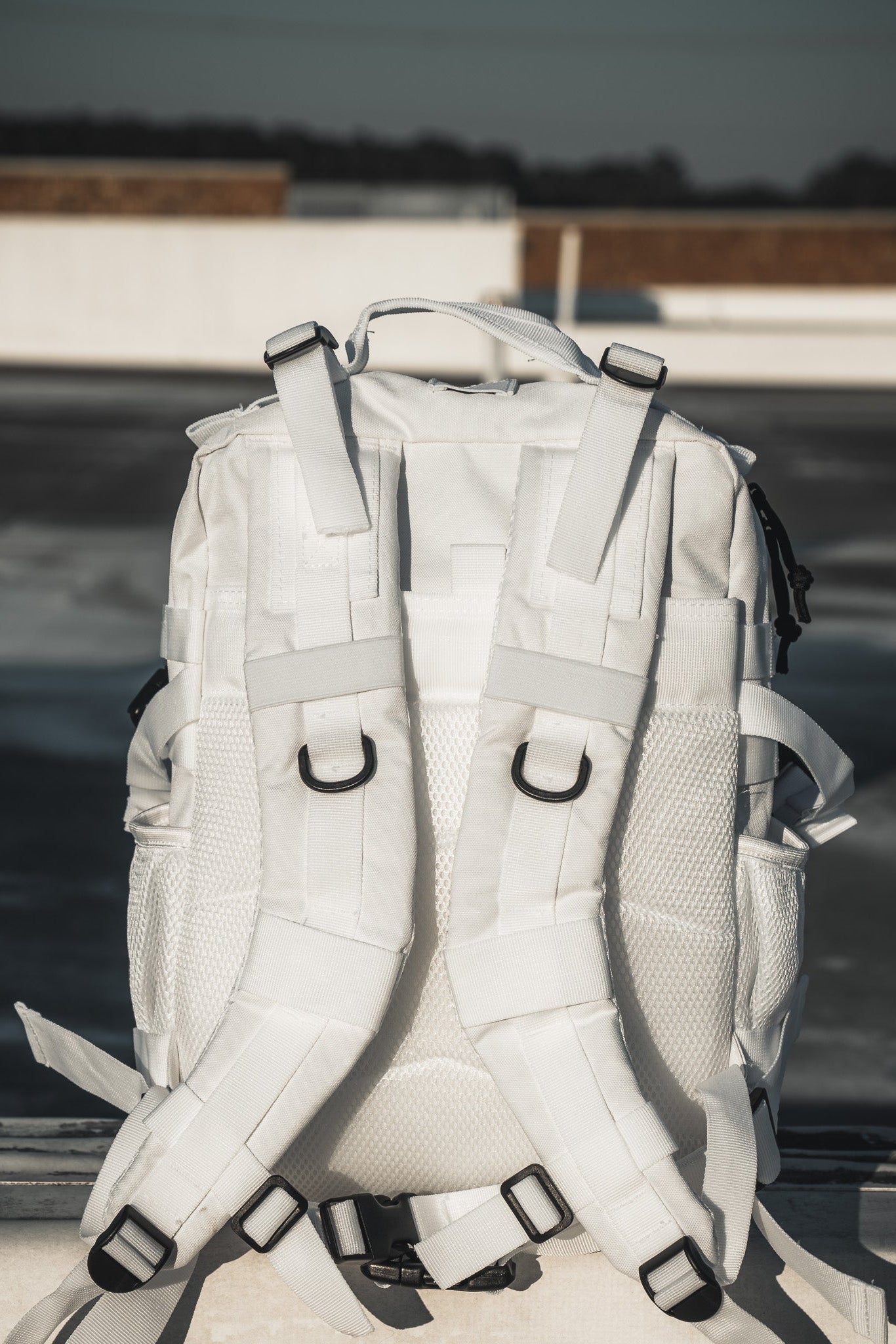 The Born United Private Label Storm Trooper Tac-Pac - 25L, with multiple straps and mesh pockets, rests on concrete outdoors, its striking white color channeling Star Wars energy against a blurred urban rooftop backdrop.