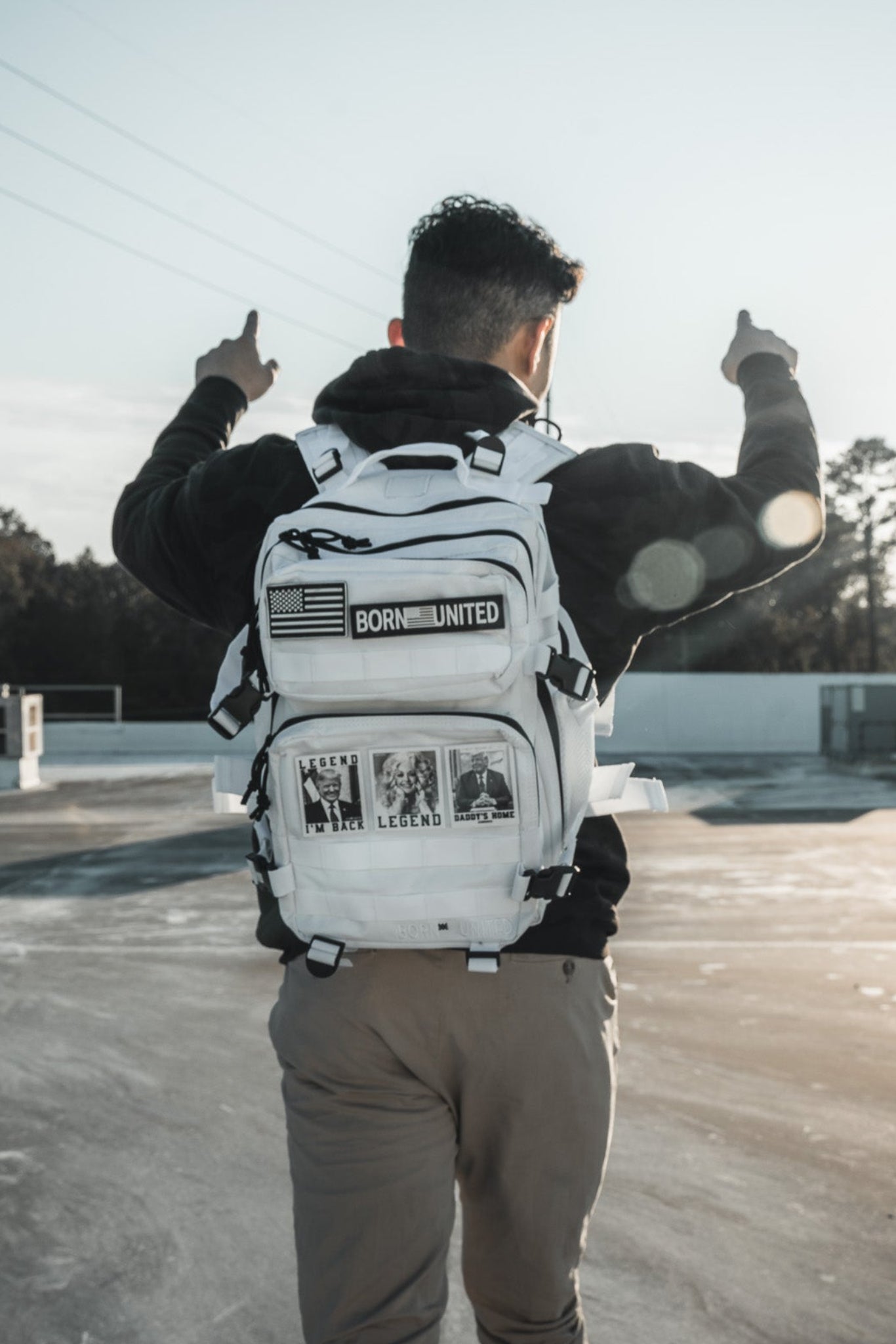 A person stands on a rooftop, arms raised, wearing a Born United Private Label Storm Trooper Tac-Pac - 25L backpack adorned with patches and photo prints. The scene features trees and bright sunlight in the background.