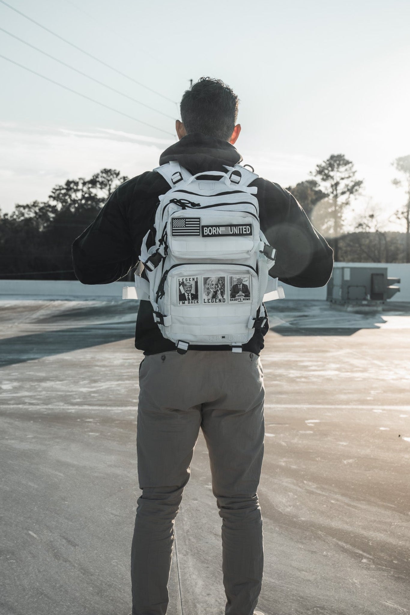 A person stands outdoors on concrete, wearing the Born United Private Label Storm Trooper Tac-Pac - 25L, a Star Wars-inspired white backpack with black patches and graphics; trees and utility lines appear in the background at sunrise or sunset.