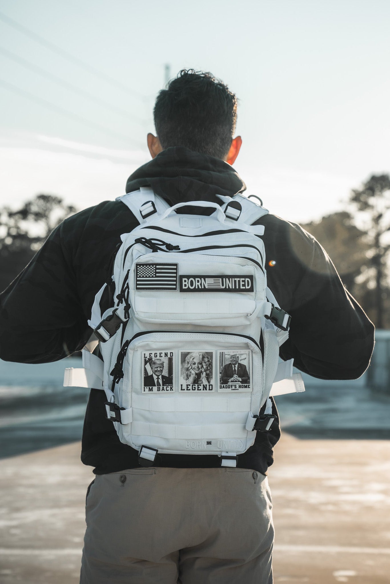 A person with their back to the camera wears a black jacket and light pants, carrying the Born United Private Label Storm Trooper Tac-Pac - 25L backpack outdoors. Trees and a clear sky are visible in the background.