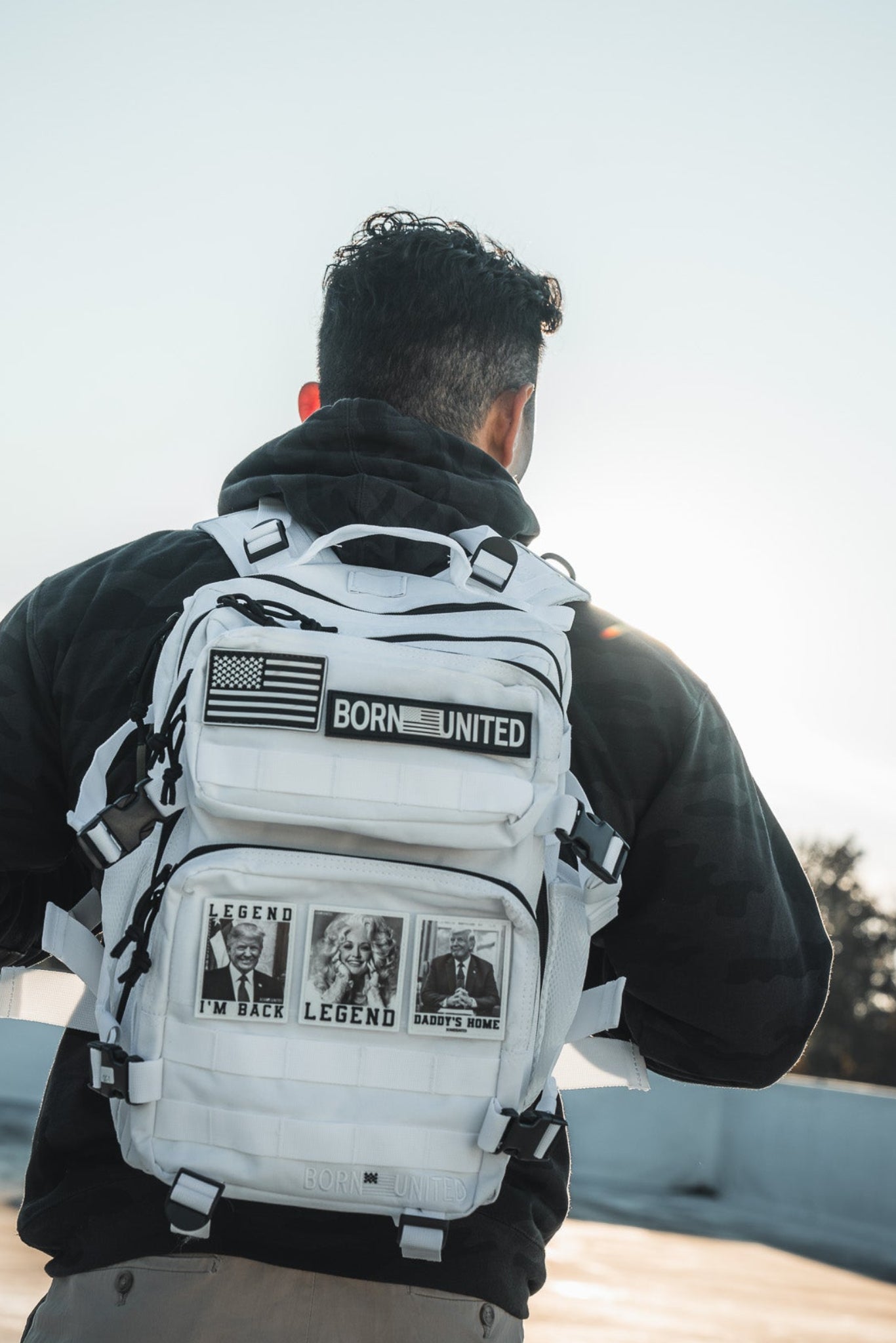A person with short dark hair, in a black hoodie and beige pants, stands outdoors at sunset with their back to the camera, carrying the Born United Private Label Storm Trooper Tac-Pac - 25L backpack decorated with patches and photos.