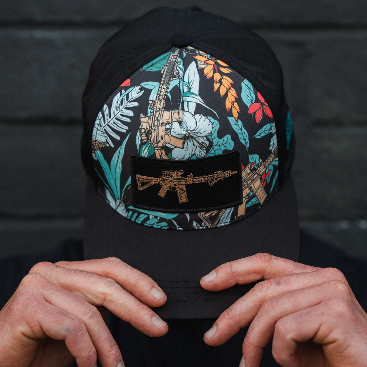 A person wearing a Tango Charlie Apparel Tactical Tropics Hydro Snapback Hat, featuring floral camouflage and an embroidered rifle design, adjusts the snapback closure against a dark textured wall in the background.