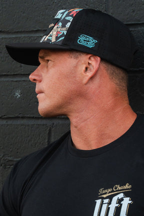 A man in a Tango Charlie Apparel Tactical Tropics Hydro snapback hat features a colorful design and T-shirt, looking to the side while standing against a dark brick wall.