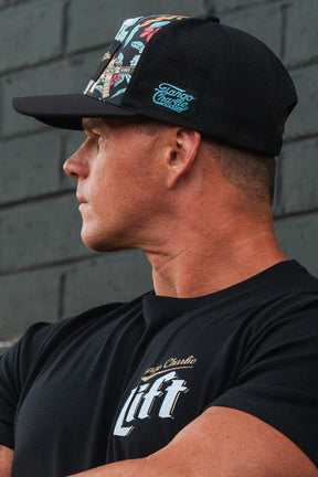 A person wearing a Tactical Tropics Hydro - Snapback Hat by Tango Charlie Apparel and a Tactical Tropics T-shirt stands in profile against a gray brick wall, looking to the side.