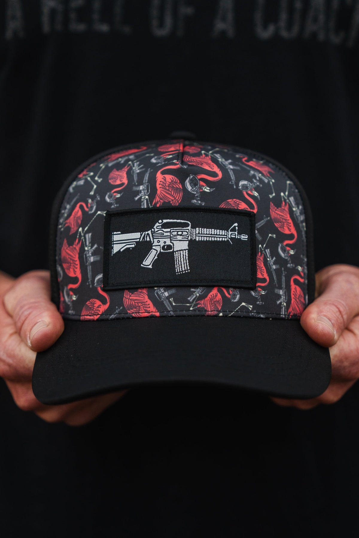 An individual holds a Tango Charlie Apparel Tactimingo Hydro snapback hat, featuring a sweat-resistant red and gray design with guns and buffalo, centered by a patch illustrating a white rifle.