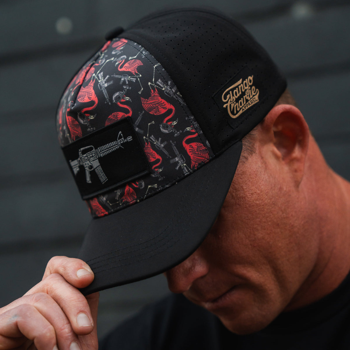 A man in a Tango Charlie Apparel Tactimingo Hydro snapback hat, featuring a sweat-resistant black fabric, red and gray flamingo and rifle pattern, tilts his head down. The cap includes a small brown patch with text on the side.