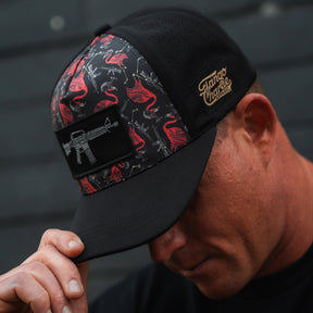 A man in a Tango Charlie Apparel Tactimingo Hydro snapback hat, featuring a sweat-resistant black fabric, red and gray flamingo and rifle pattern, tilts his head down. The cap includes a small brown patch with text on the side.