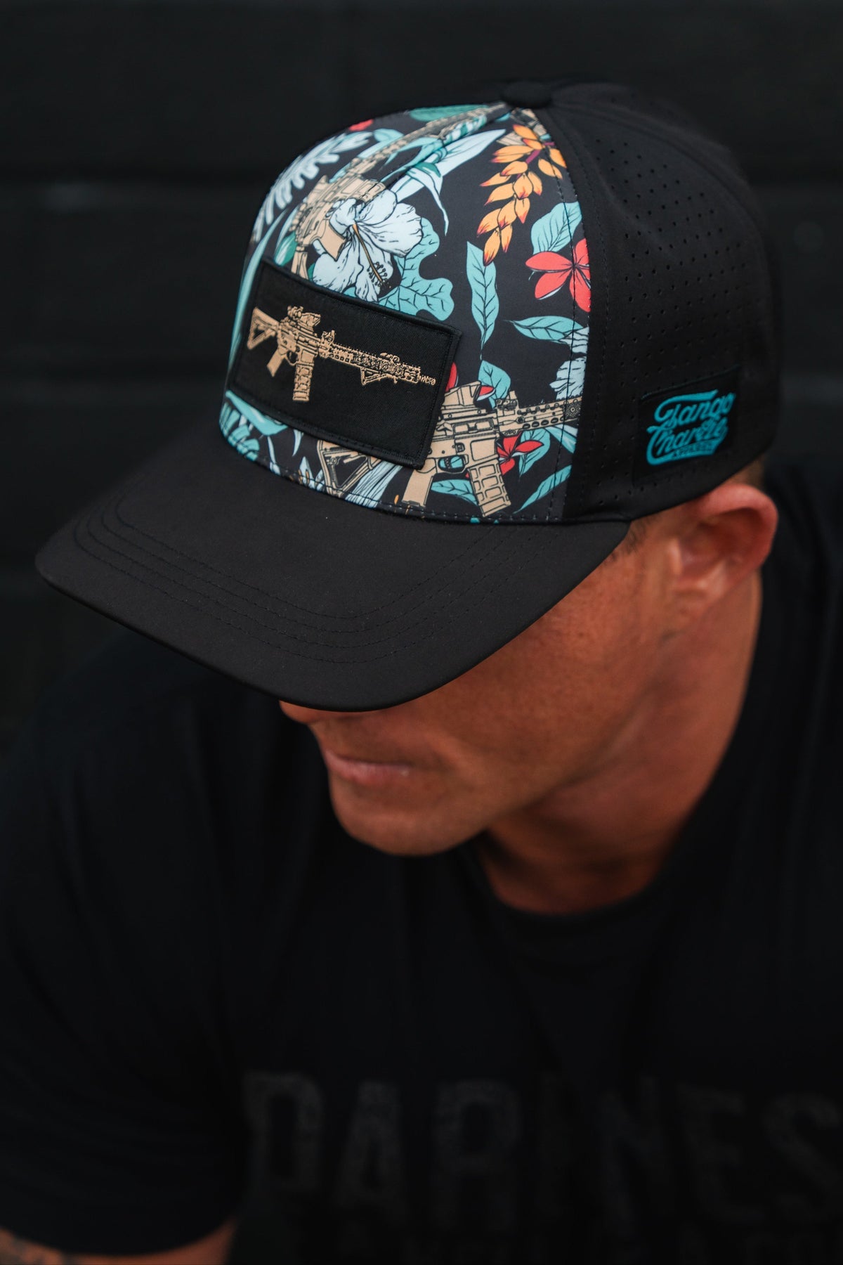 Wearing the Tactical Tropics Hydro Snapback Hat by Tango Charlie Apparel, featuring a vibrant floral pattern and gun patch, the individual in a dark T-shirt completes their look with this stylish black cap. Their face remains unseen.