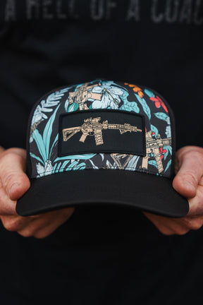 Someone is holding a Tactical Tropics Hydro - Snapback Hat from Tango Charlie Apparel. This hat features a dark, vibrant floral pattern with an embroidered rifle patch on the front and includes a snapback enclosure.