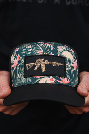 A person holds the Tango Charlie Apparel Aloha AR Hydro Snapback Hat, showcasing pink and white floral patterns over a dark background with a rifle patch.
