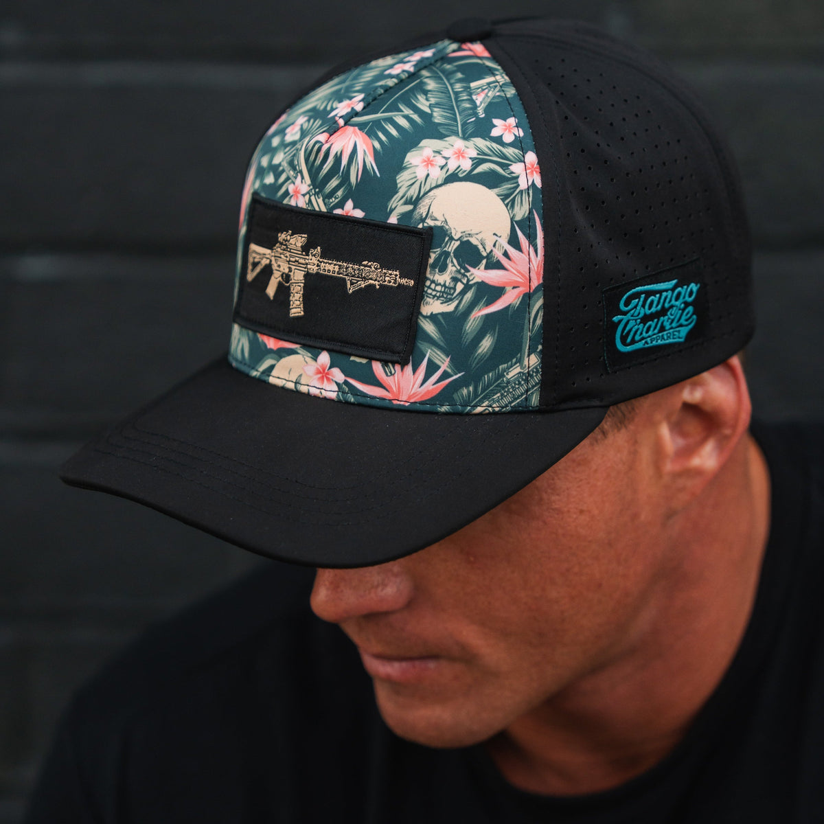 A man dons a black Tango Charlie Apparel Aloha AR Hydro Snapback Hat with a floral and skull design, an embroidered rifle on the front, side perforations for breathability, and a blue logo patch while standing against a dark wall.